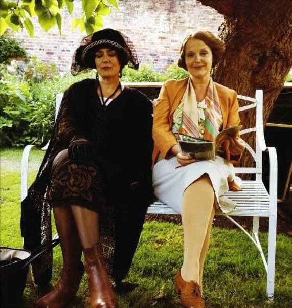 Anna Chancellor and Miranda Richardson  sitting on a white garden bench under a tree, one dressed in vintage black with a large hat, the other in a modern beige blazer, holding a magazine.