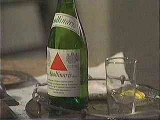 A glass bottle of Apollinaris mineral water tipping over, spilling water into a glass on a table.