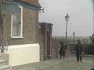 Two people walking down a cobblestone street near a brick building with street lamps along the path.