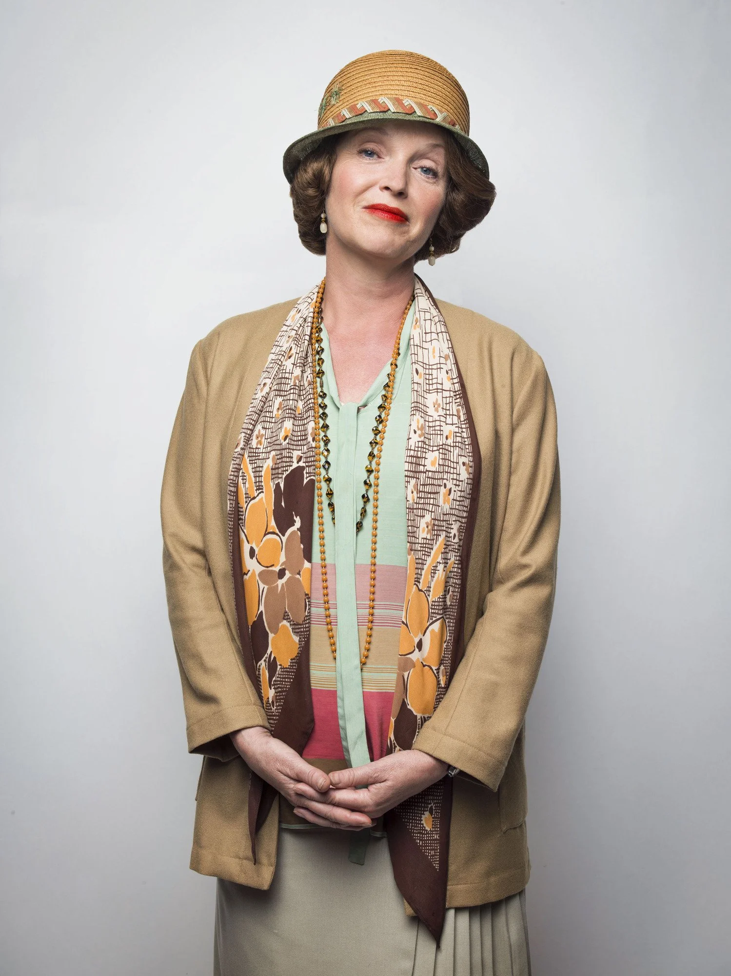 Miranda Richardson wearing a tan coat, colorful scarf, and a multicolored straw hat with a decorative band, standing against a plain background.
