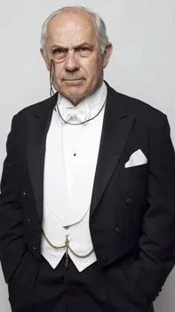 A distinguished elderly man in a black tuxedo with a white shirt and bowtie, standing against a plain background.