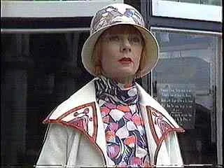 A woman with a blonde bob hairstyle wearing a wide-brimmed hat, a colorful patterned scarf, and a white coat, standing outdoors in front of a building with glass windows.