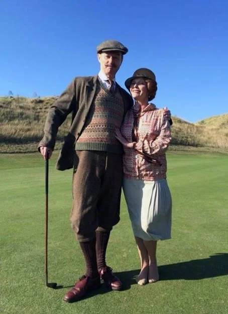 Mark Gatiss and Miranda Richardson dressed in vintage clothing standing on a golf course with green grass and hills in the background, smiling and posing for the photo.
