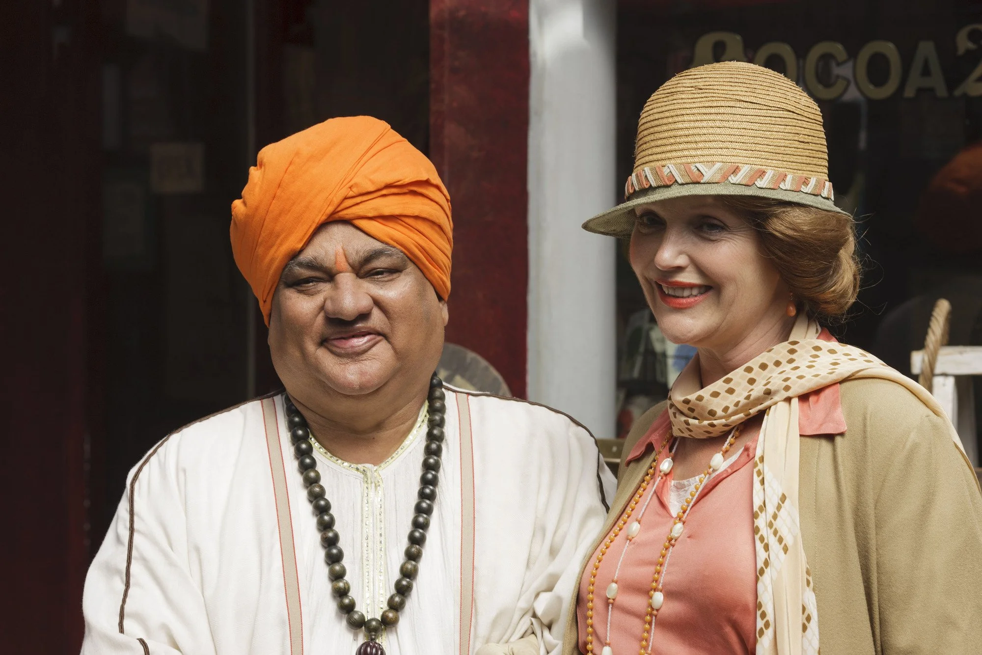 The Guru and Mapp dressed in vintage attire standing outdoors, one wearing an orange turban and traditional Indian clothing, the other wearing a straw hat and polka-dot scarf, smiling.