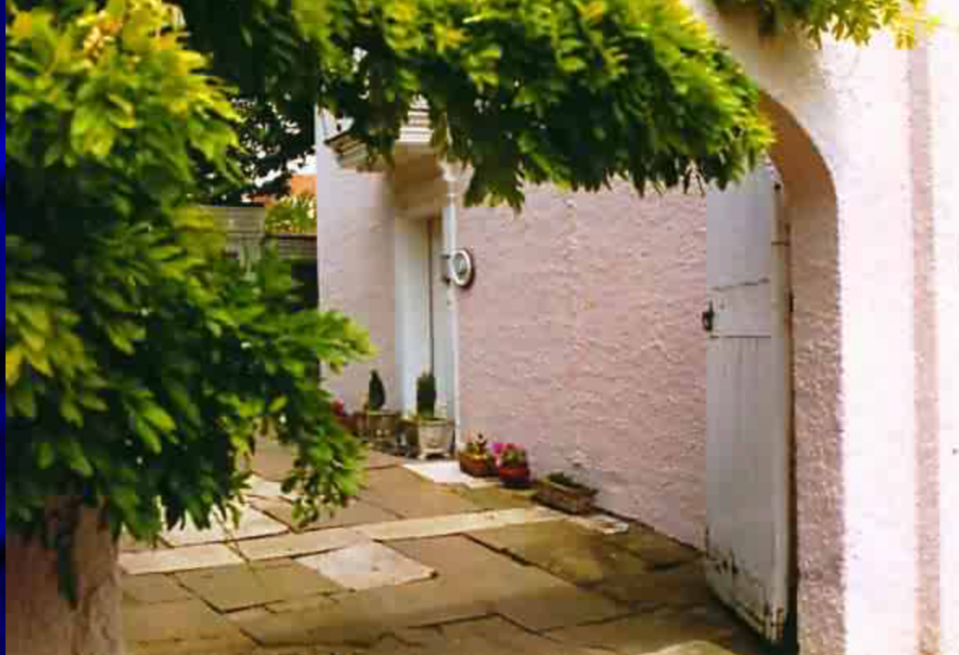 A quaint country pathway with stone paving leading to a light pink wall with a white wooden door, shaded by a large green tree, and decorative potted plants along the side.