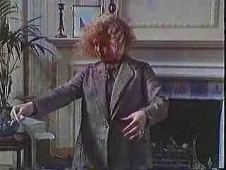 Woman with curly red hair reaching for something on a desk in a living room with a fireplace and plant.