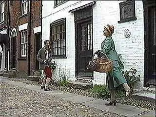 A woman with a hat and green coat carrying a basket approaches two children, one holding a book, on a street with old buildings.