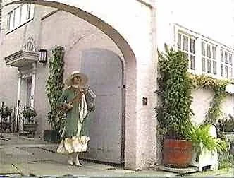 A woman with a straw hat and light-colored clothing walking near a white building with plants and an archway.