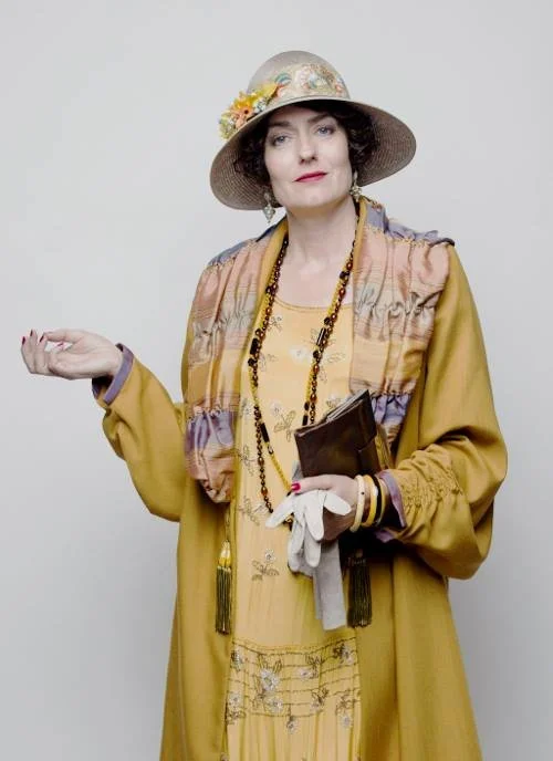 Anna Chancellor wearing a wide-brimmed hat decorated with flowers, a yellow dress, a multi-colored scarf, and layered jewelry. She holds a small clutch purse, white gloves, and a pair of glasses, standing against a plain light background.