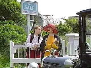 Two women standing outdoors near a white picket fence, with one holding a red umbrella and a teddy bear, appearing to converse.