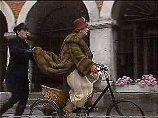 A woman riding a bicycle with a basket, being followed by a person pushing the bike from behind in an urban setting.
