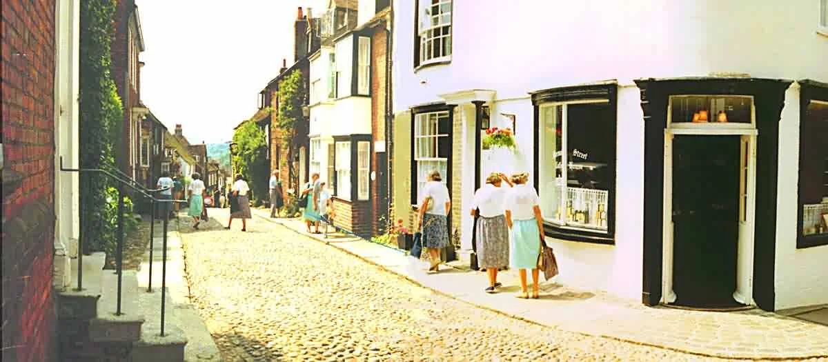 People walking down a cobblestone street lined with shops and buildings on a sunny day.