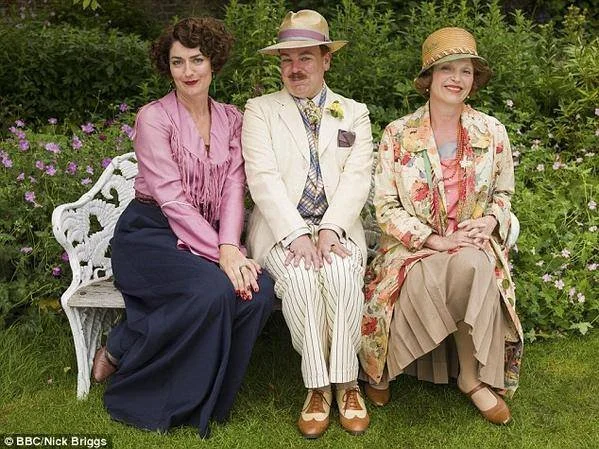 Steve Pemberton, Anna Chancellor and Miranda Richardson  sitting on a white bench outdoors, dressed in vintage clothing, surrounded by green foliage and flowers.