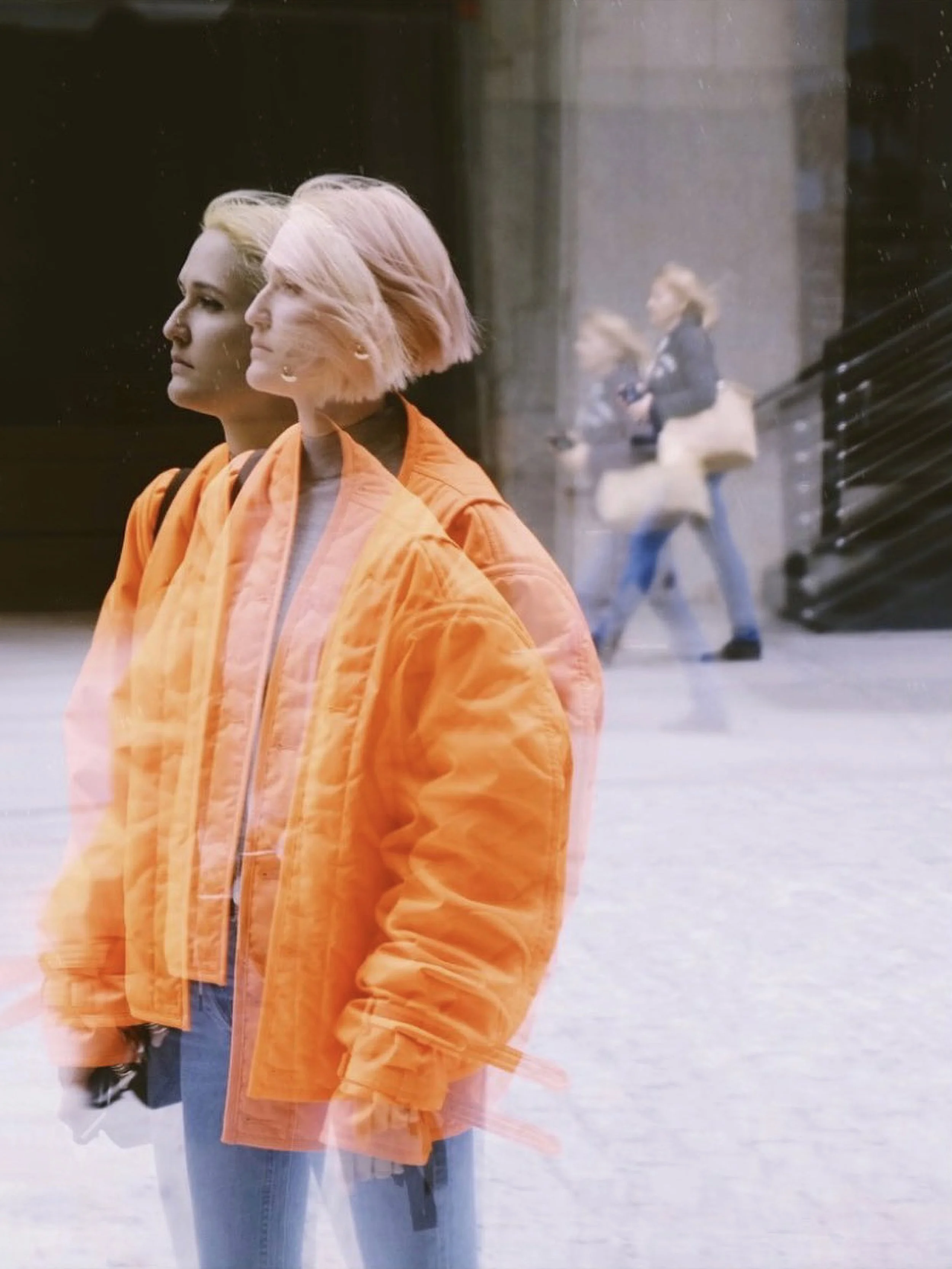 Woman in an orange jacket walking past a reflective storefront, with her reflection and a blurred person visible in the glass.