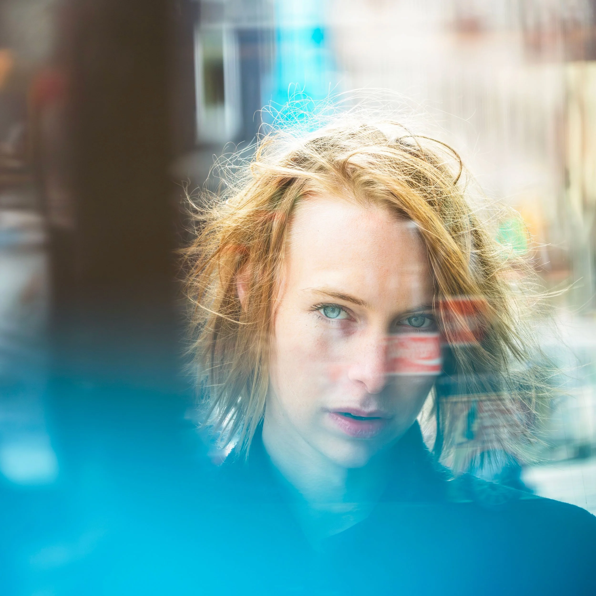Close-up portrait of a woman with tousled blonde hair and blue eyes behind a reflective window