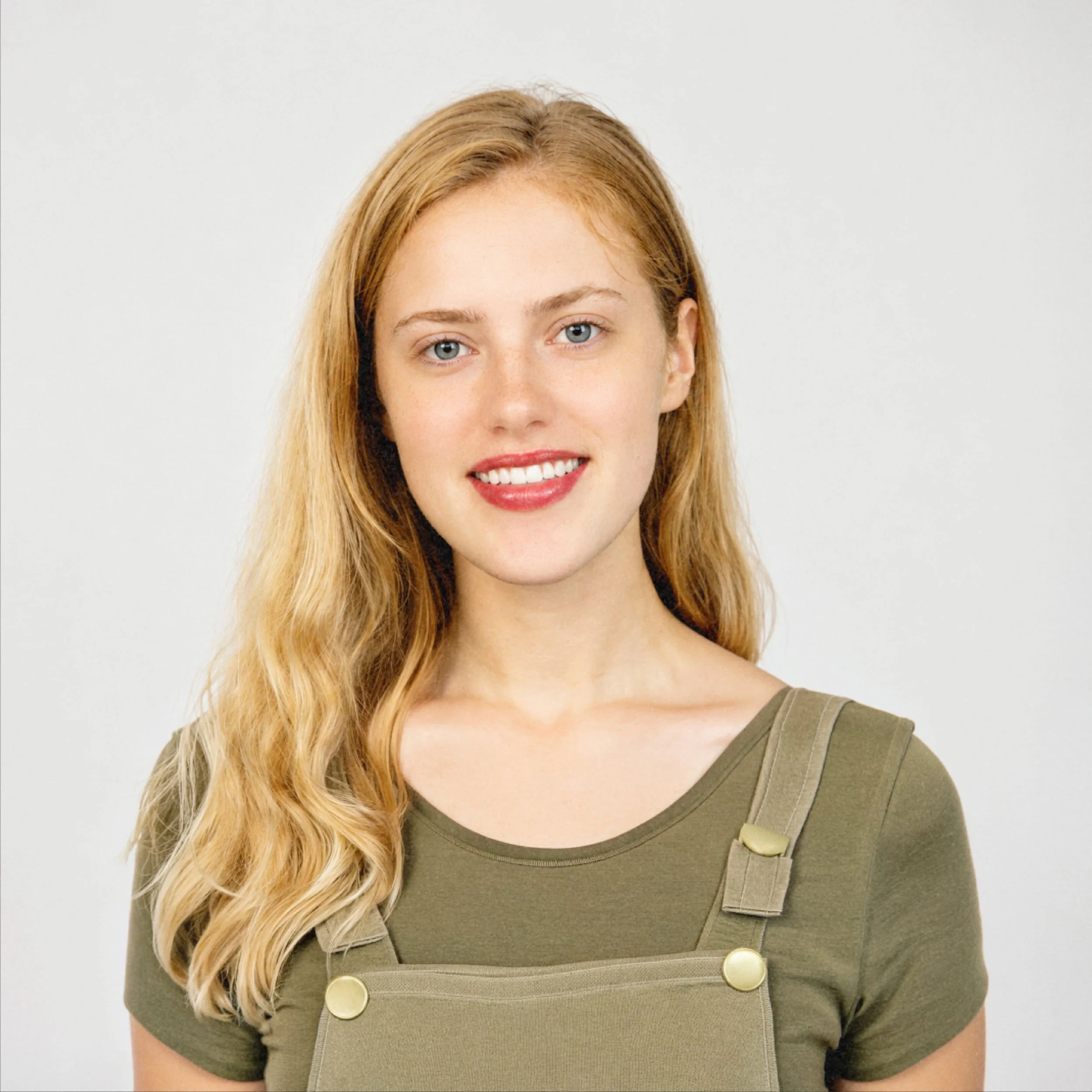Smiling blonde woman wearing an olive green T-shirt and overalls against a white background