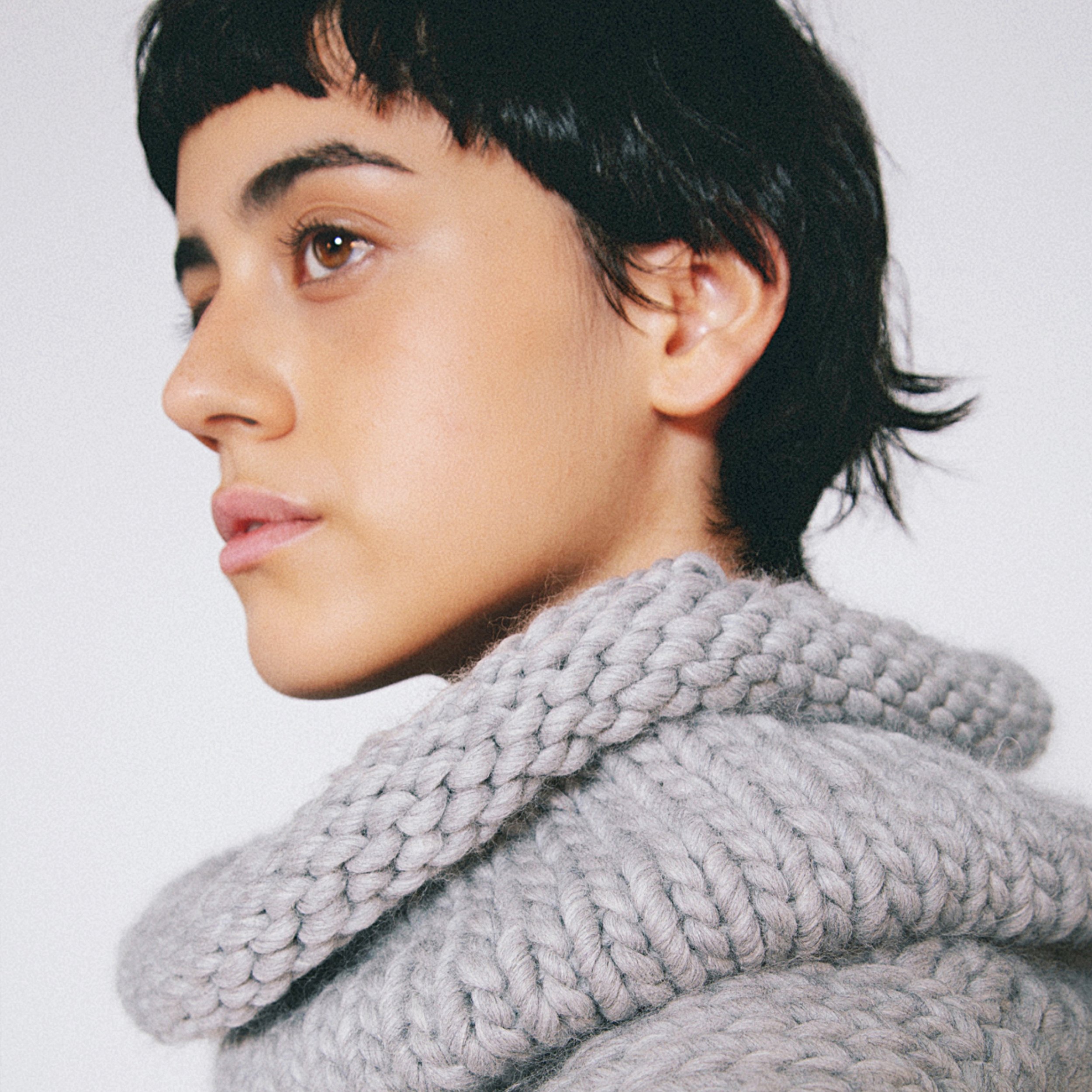 Close-up portrait of a person with short dark hair and brown eyes wearing a chunky gray knit sweater