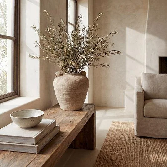 A rustic wooden table with a ceramic vase filled with dried branches, a stack of books, and a ceramic bowl, next to a large window with natural light, in a neutral-toned living room