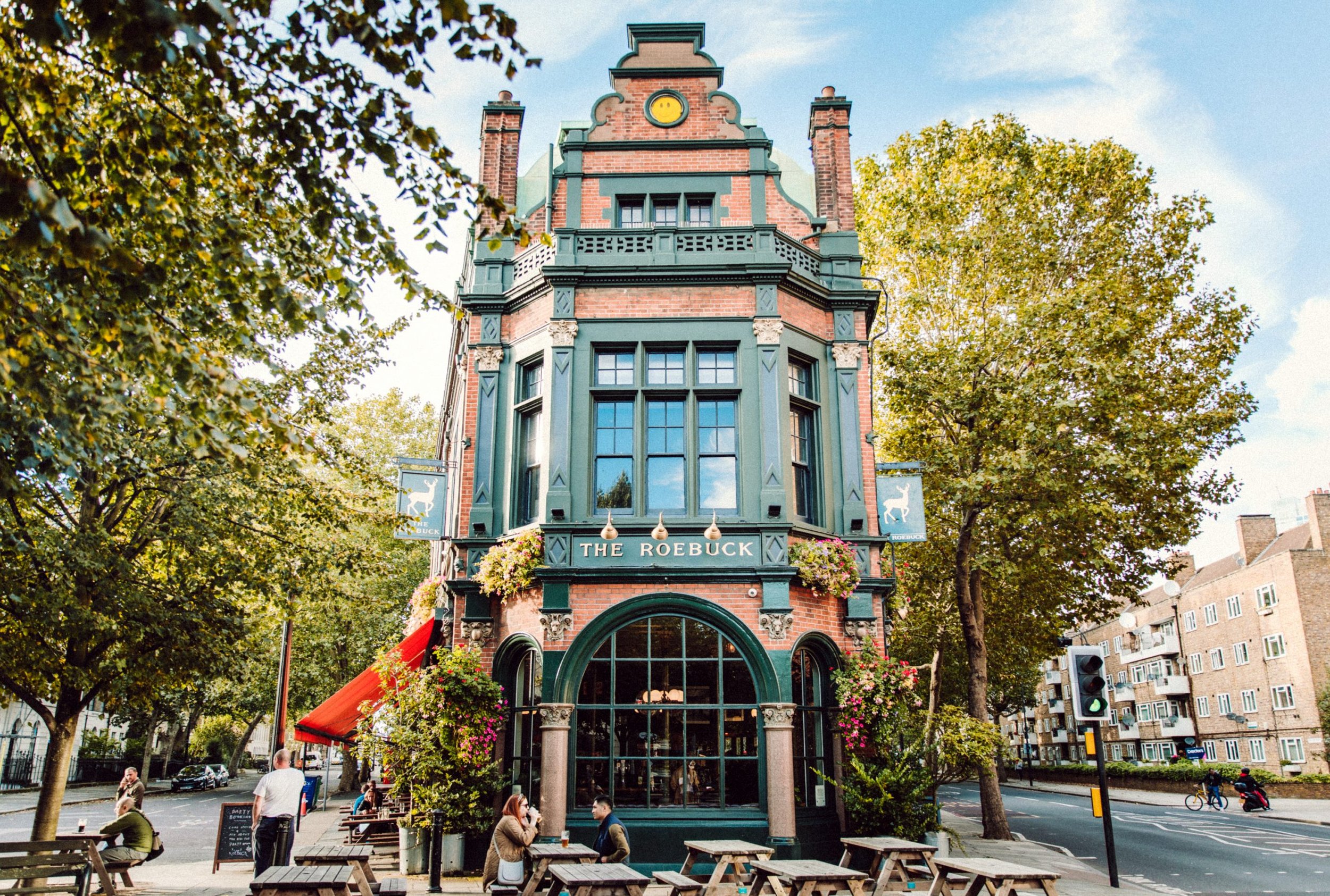A historic building with green accents and a sign reading 'The Roebuck' on the front; outdoor seating area with wooden tables and people sitting, surrounded by trees and city buildings.