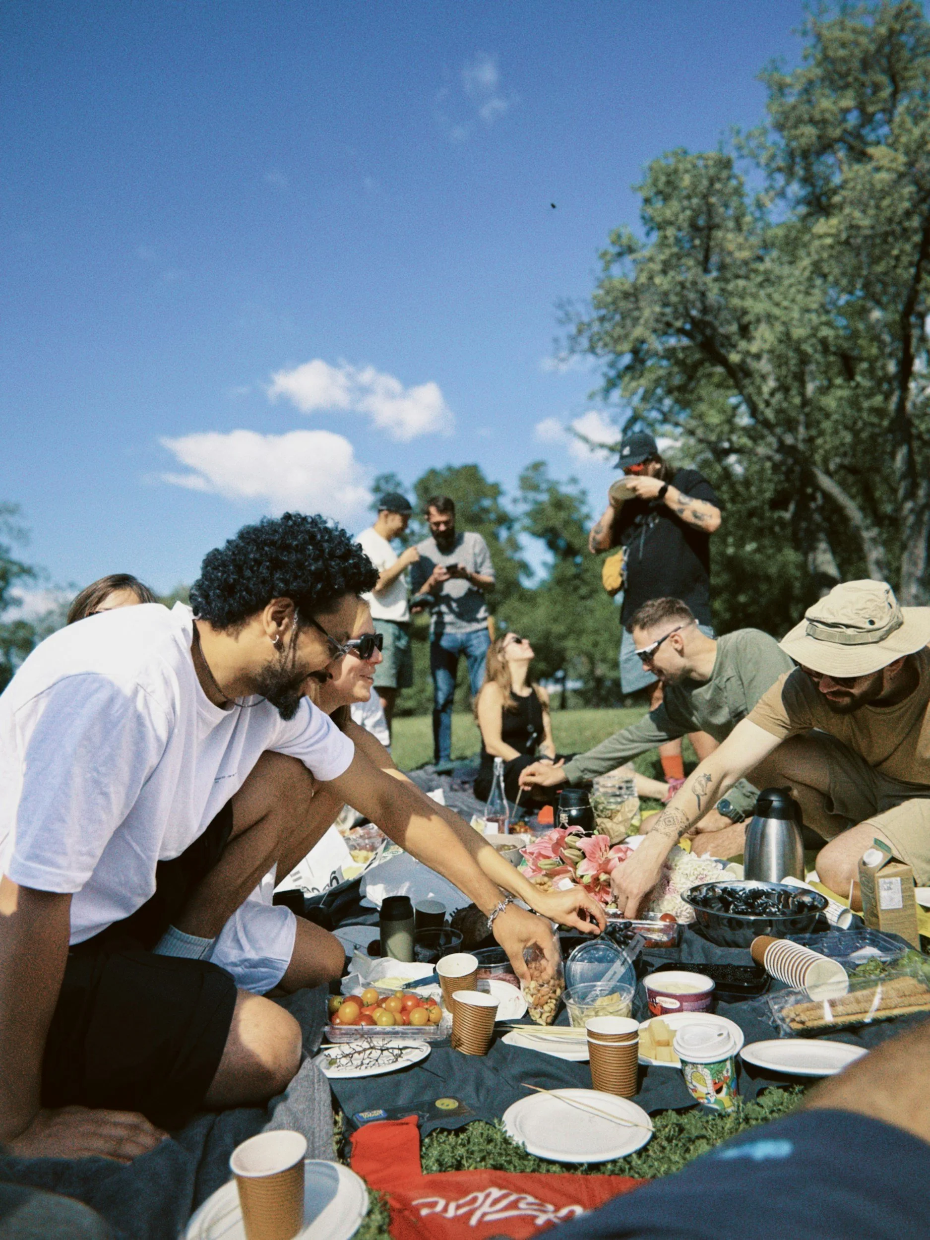 Group of people gathered outdoors during daytime, participating in an event with food and drinks on a table, under a blue sky with trees in the background.