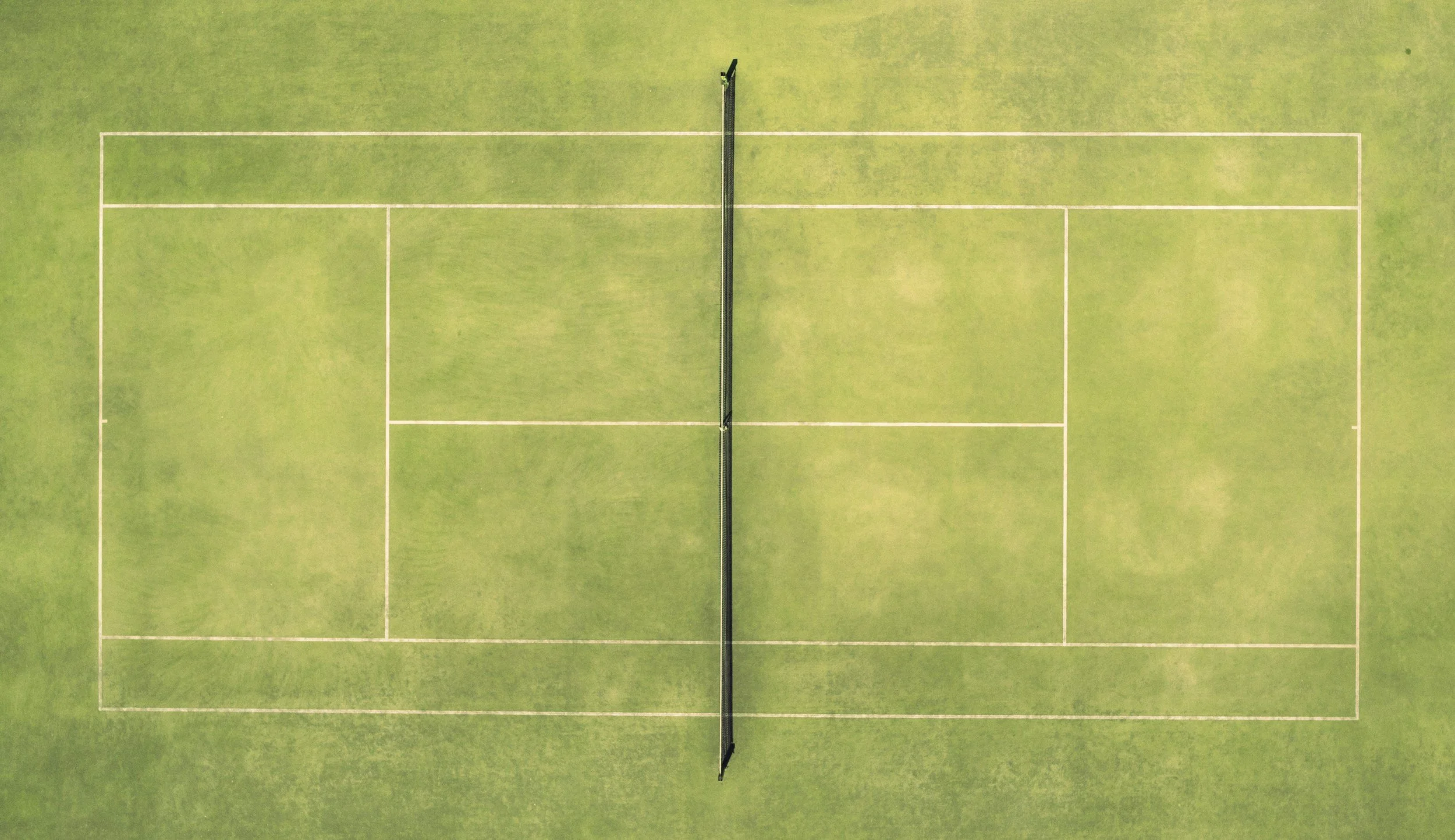 Top aerial view of a tennis court with a net in the center and white boundary lines on green surface.