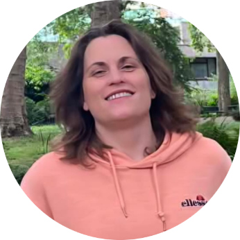 A woman with shoulder-length brown hair, smiling, wearing a peach-colored hoodie in a green outdoor park setting.