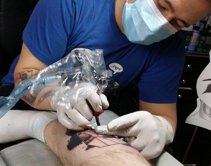 Tattoo artist wearing a face mask and gloves tattooing a geometric design on a client’s leg