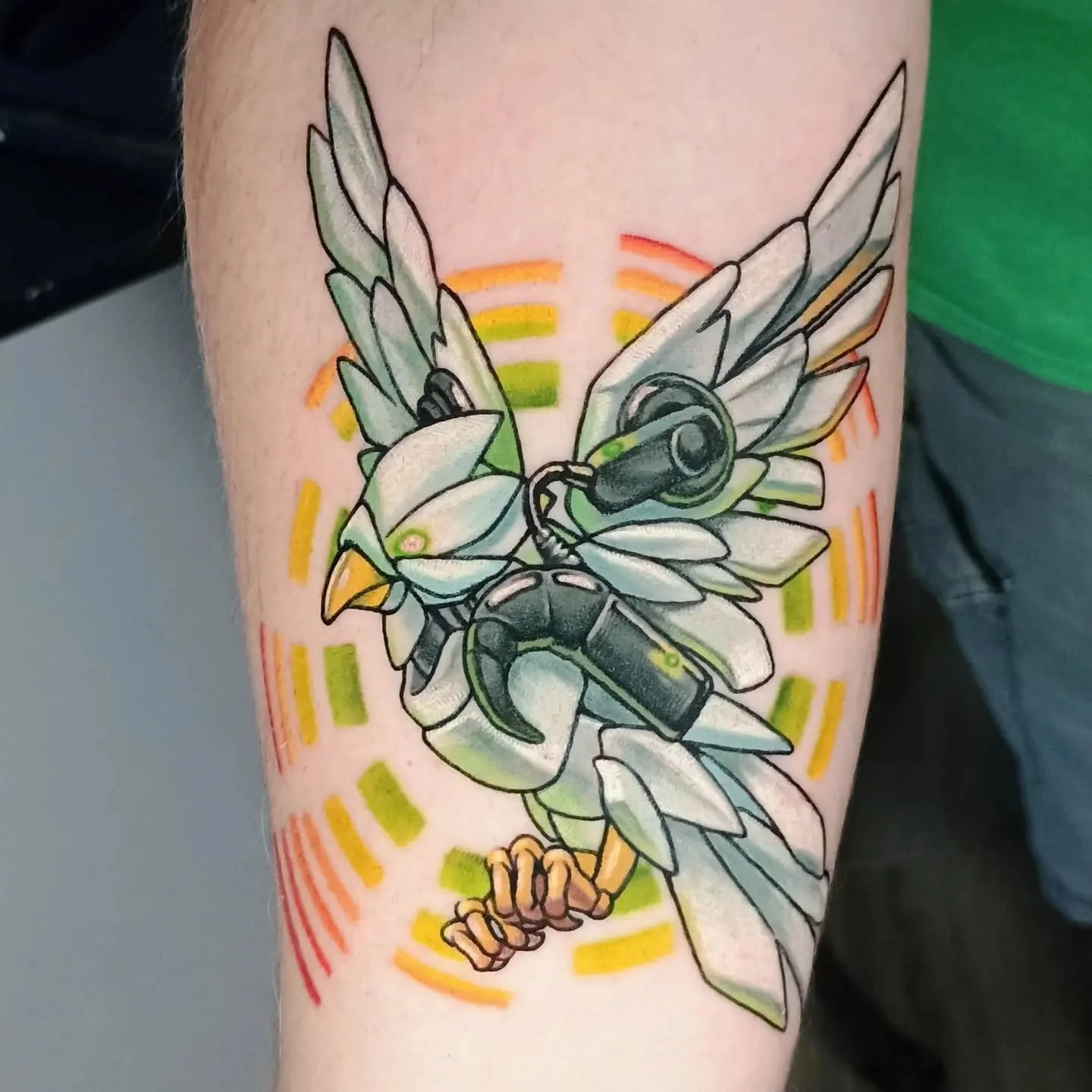 Colorful tattoo of two winged cartoon creatures with mechanical details and orange, yellow, and green geometric accents on a person’s leg