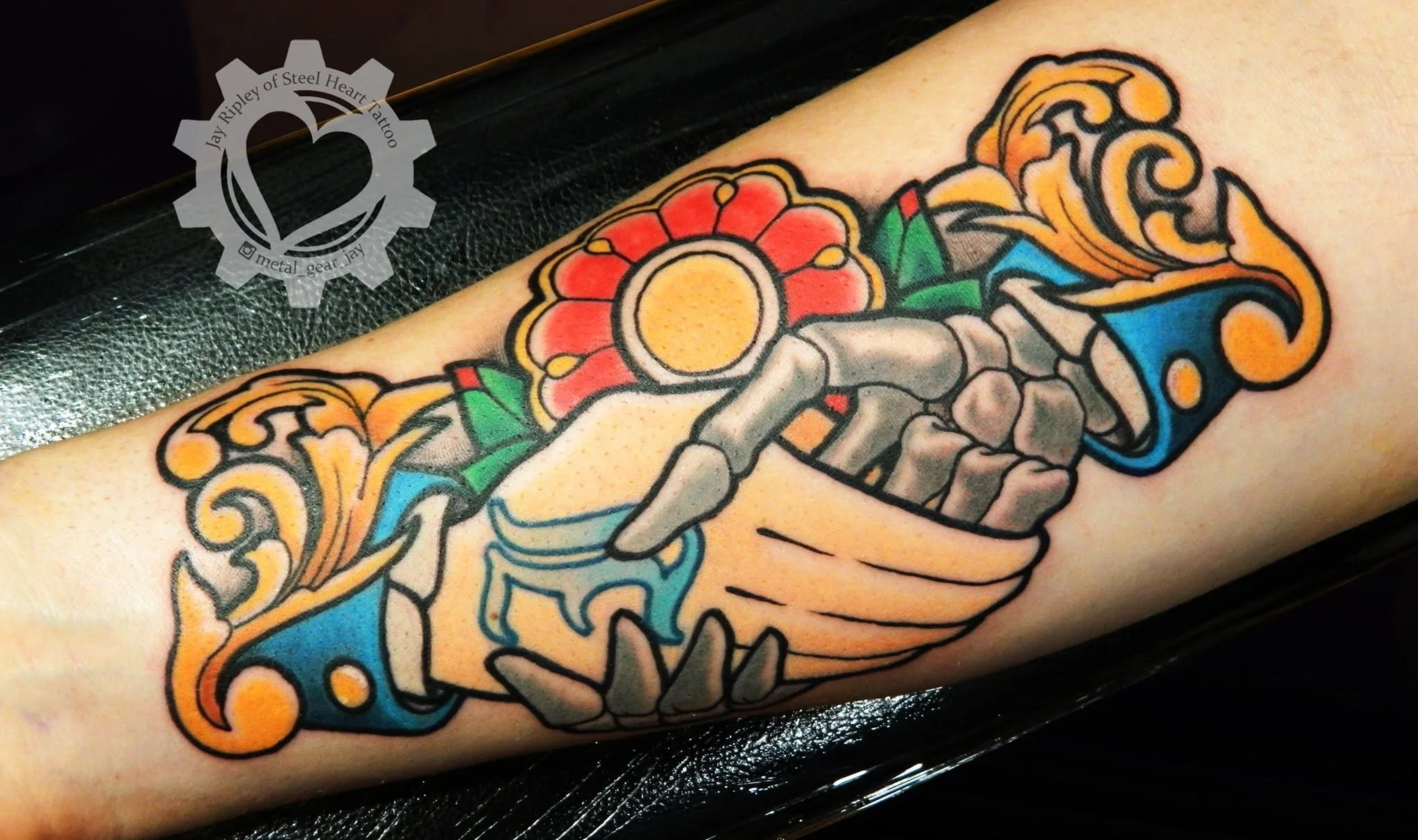 Colorful traditional-style tattoo featuring a skeleton hand holding a red flower with blue and orange ornamental scrollwork