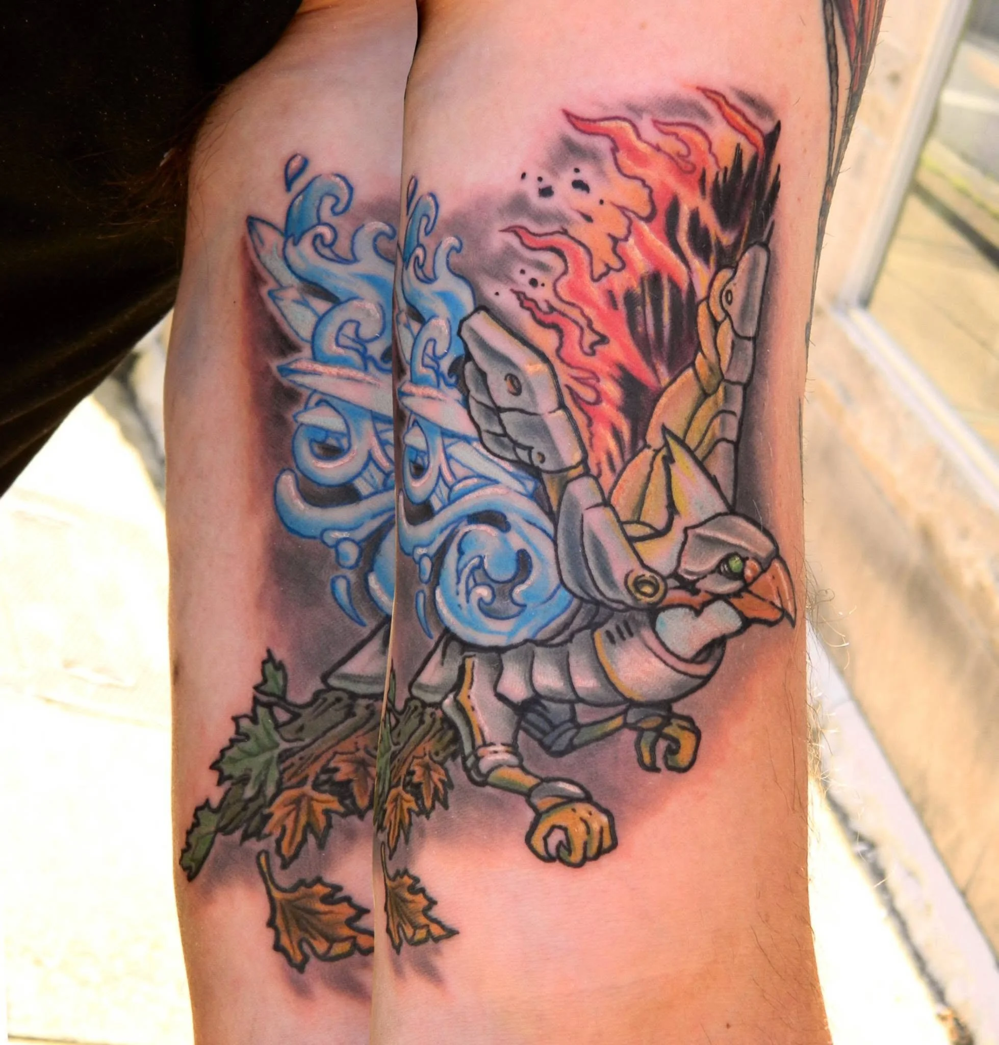 Colorful tattoo featuring a cartoonish armored bird with an orange beak, blue swirling flames, red flames, and autumn leaves.