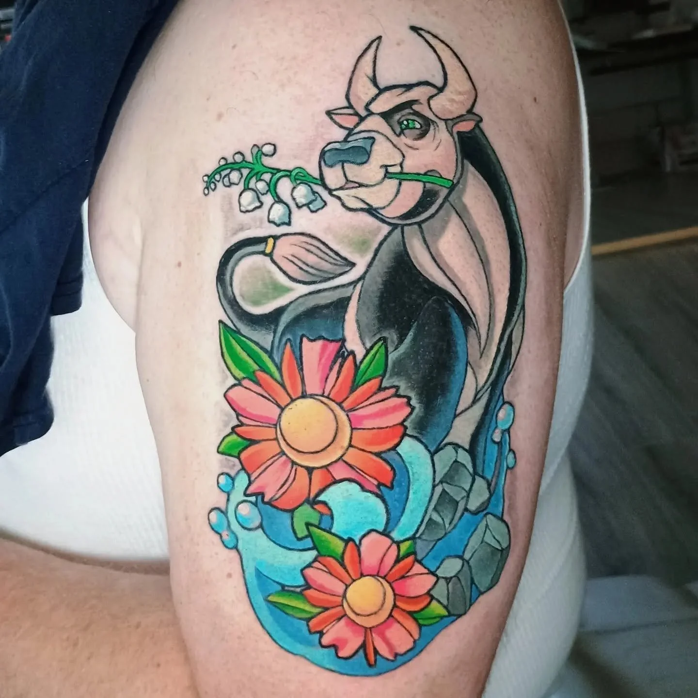 Colorful tattoo of a horned dragon holding a lily-of-the-valley sprig above flowers and curling blue water.