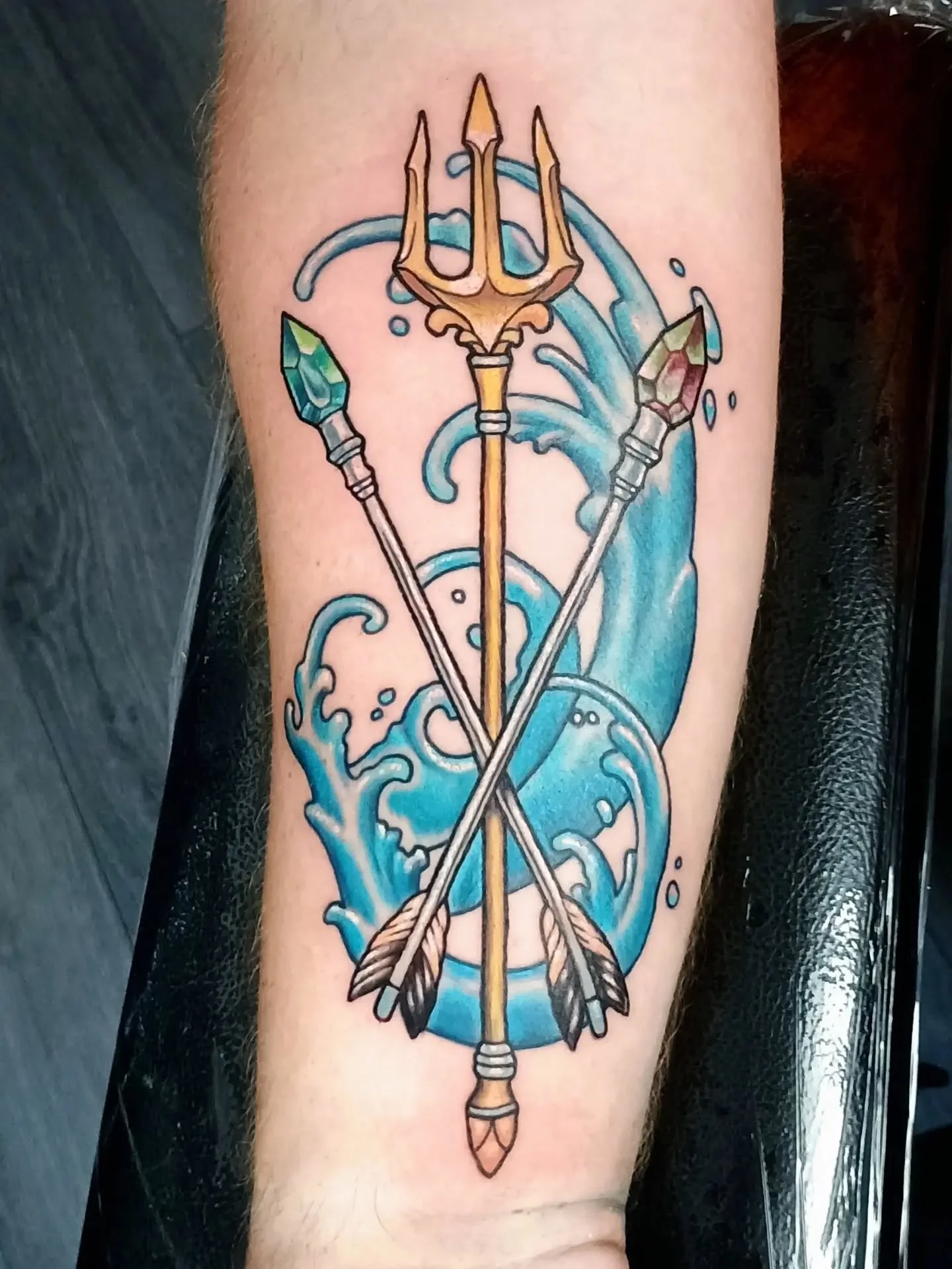 Colorful forearm tattoo featuring a golden trident, crossed crystal-tipped spears, blue ocean waves, and arrows.