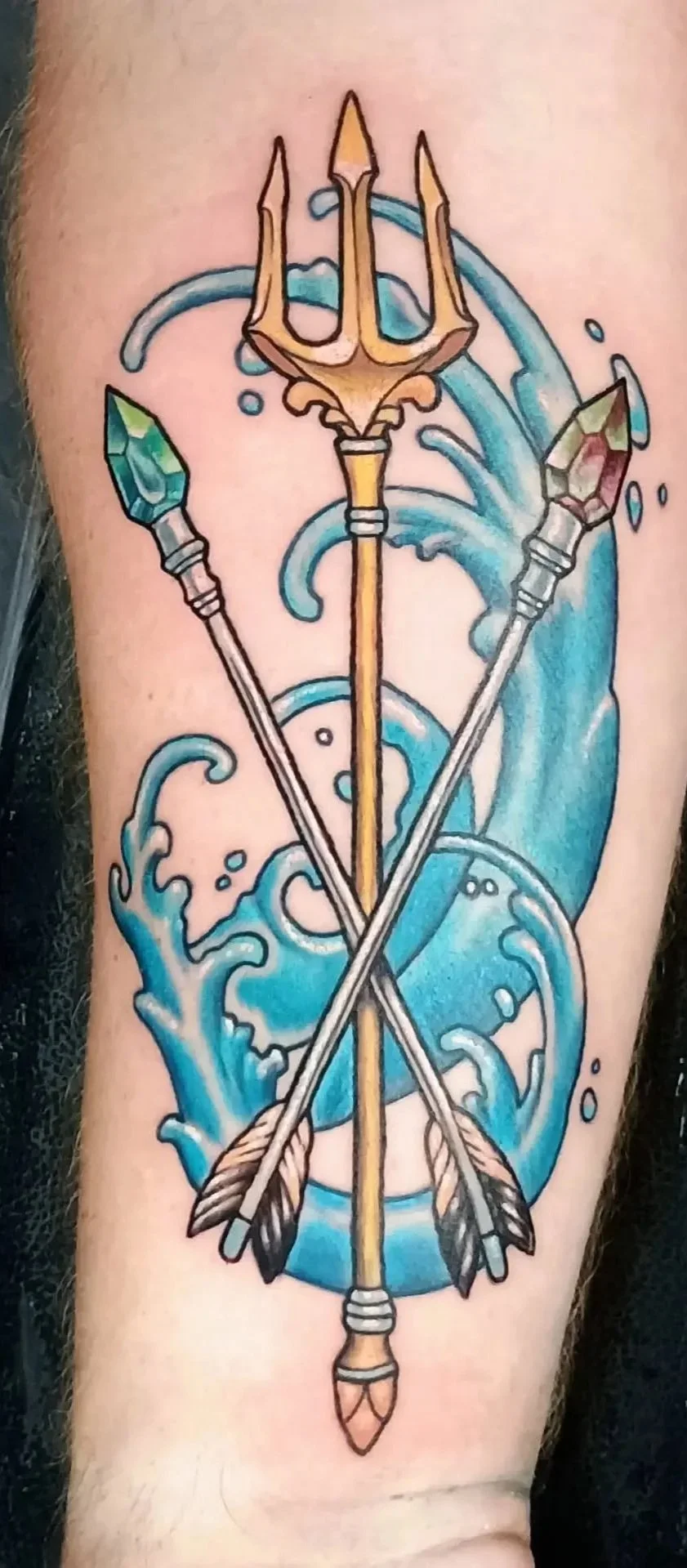 Colorful tattoo featuring a golden trident, crossed arrows with jeweled arrowheads, and stylized blue ocean waves.