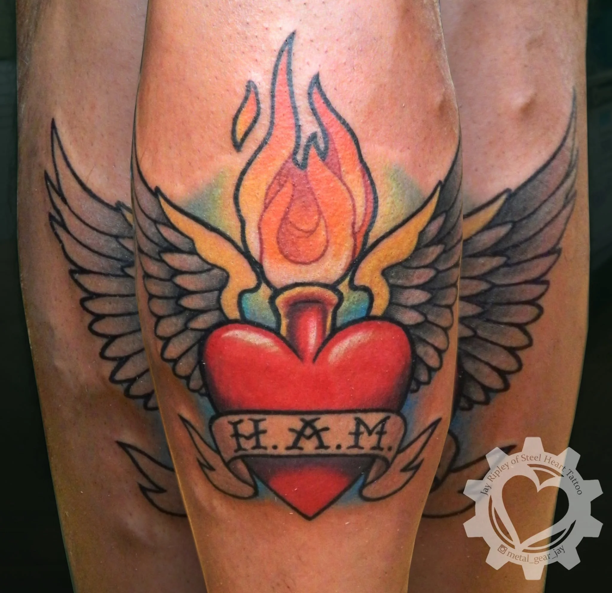 Traditional tattoo featuring a flaming red heart with wings, a crown, and a banner reading “H.I.A.M.”