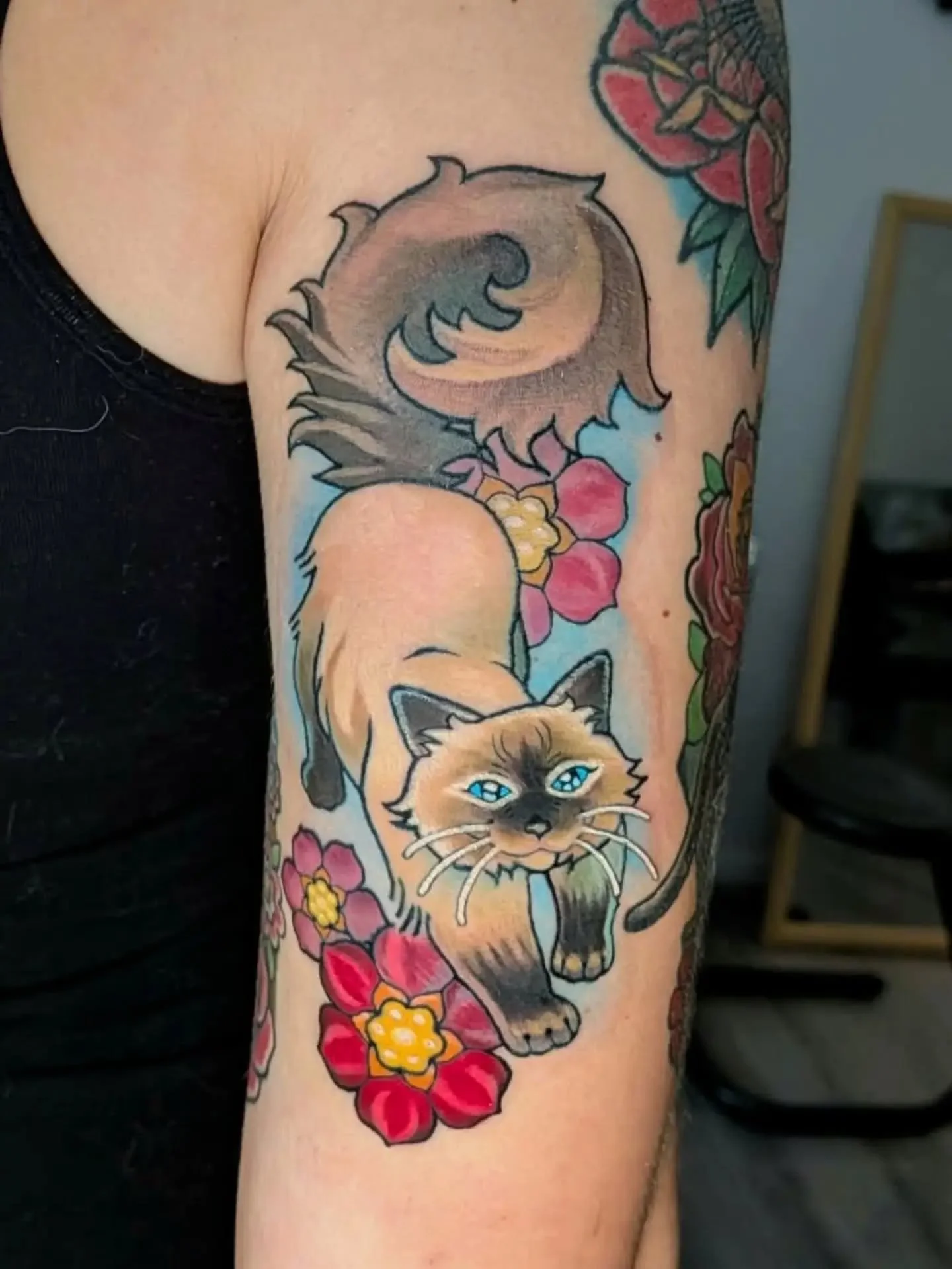 Colorful tattoo of a Siamese cat surrounded by pink flowers and a large gray bird on a person’s arm