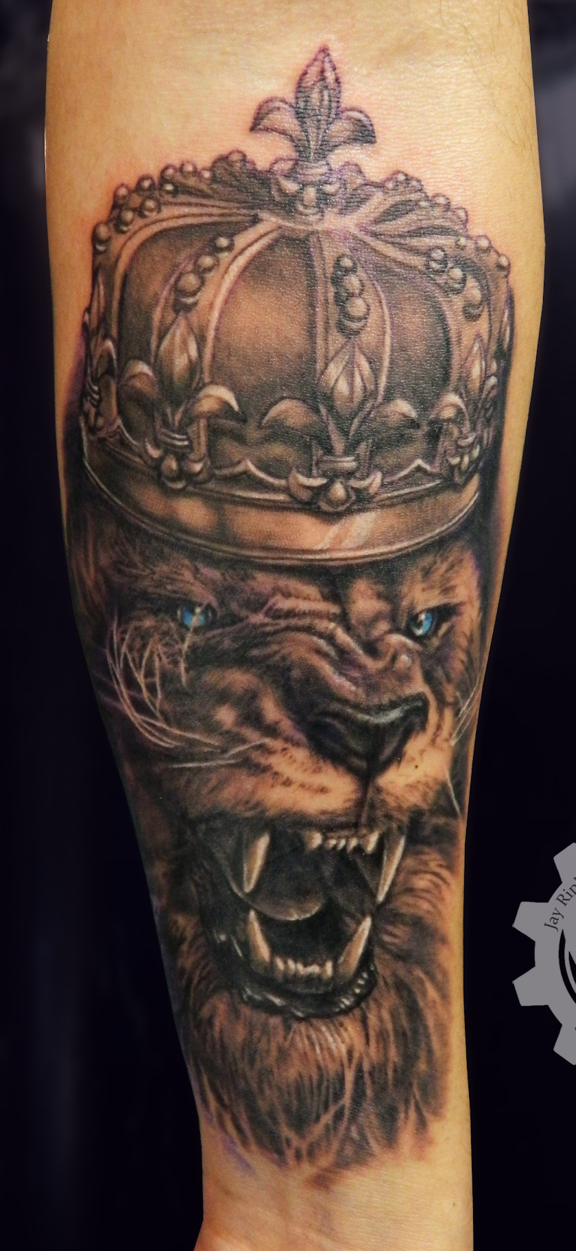 Detailed forearm tattoo of a snarling blue-eyed tiger beneath an ornate jeweled crown topped with a fleur-de-lis