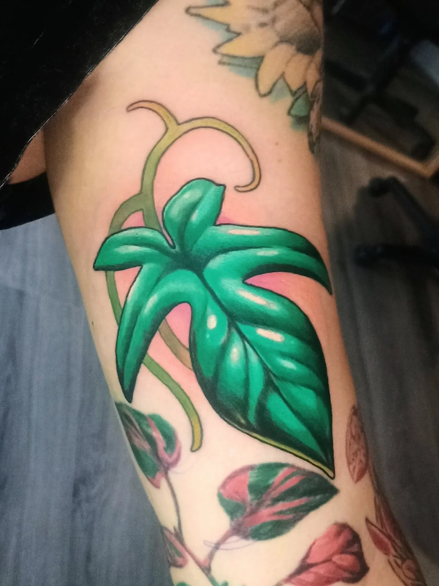 Close-up of a colorful botanical tattoo featuring large green leaves, vines, and pink flowers on an arm