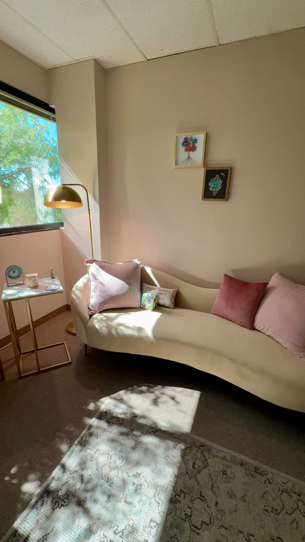 A cozy corner of a room with a beige couch decorated with pink and purple pillows, a small side table with a clock and candles, a floor lamp with a gold shade, a window with sunlight and green tree leaves outside, and two framed floral art pieces on the wall.