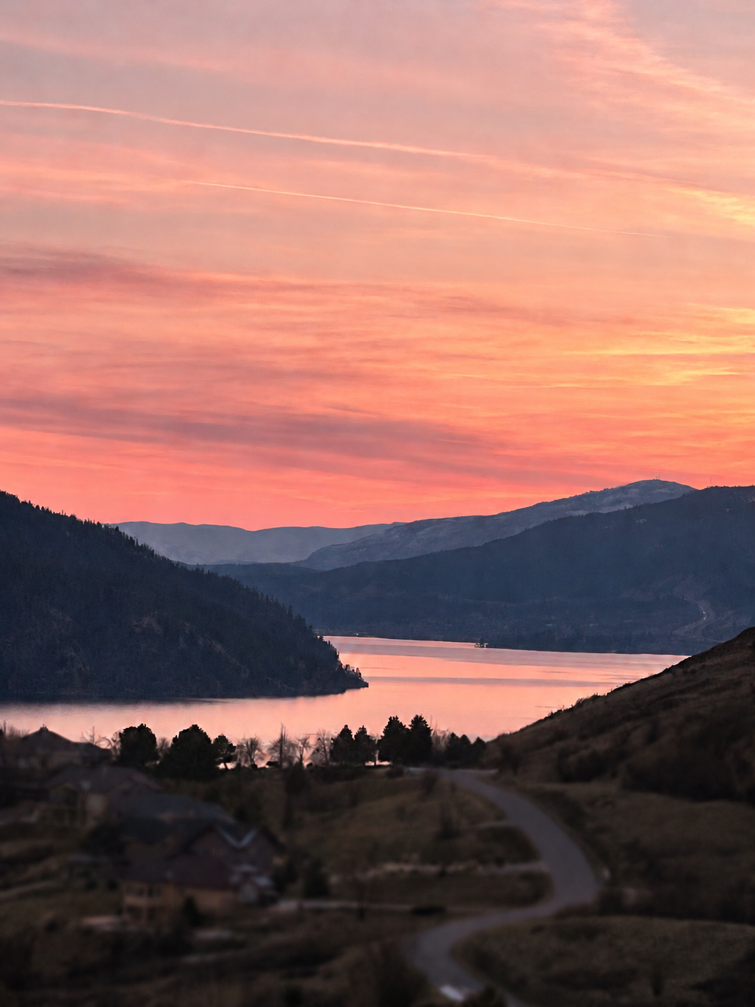 A scenic landscape of a river surrounded by rolling hills and mountains at sunset, with a pink and orange sky and a few wispy clouds.