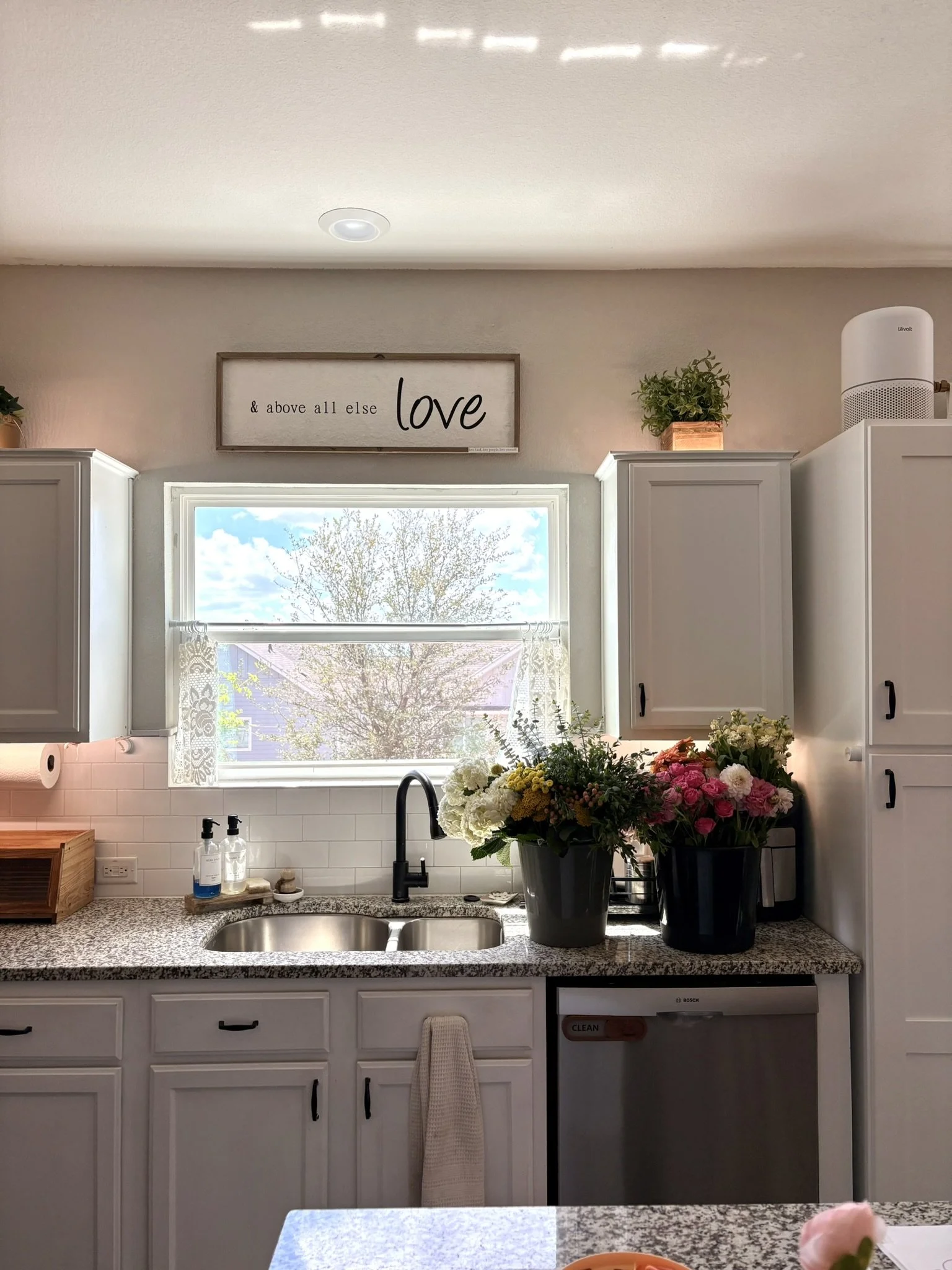 A kitchen with white cabinets, a granite countertop, and a window showing a tree outside. There are two large vases with flowers on the counter and a framed sign that reads '& above all else love' on the wall above the window. A soap dispenser and paper towels are also visible.