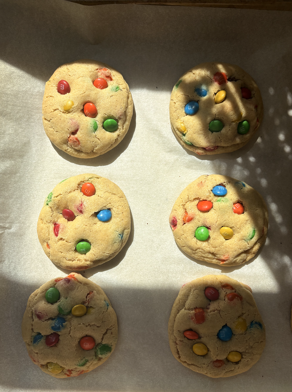 Six colorful cookies with M&M candies on top, arranged on parchment paper with sunlight and shadow patterns.