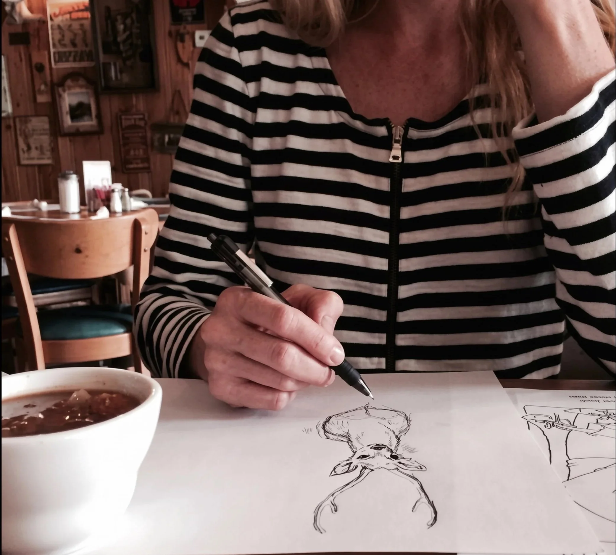 Artist-woman in a black-and-white striped shirt drawing  a deer with a pen at a coffee shop table beside a bowl of soup