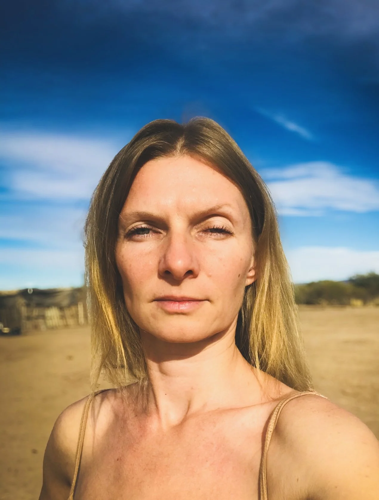 A woman artist with long blonde hair taking a selfie outside in front of a clear blue sky with wispy clouds.