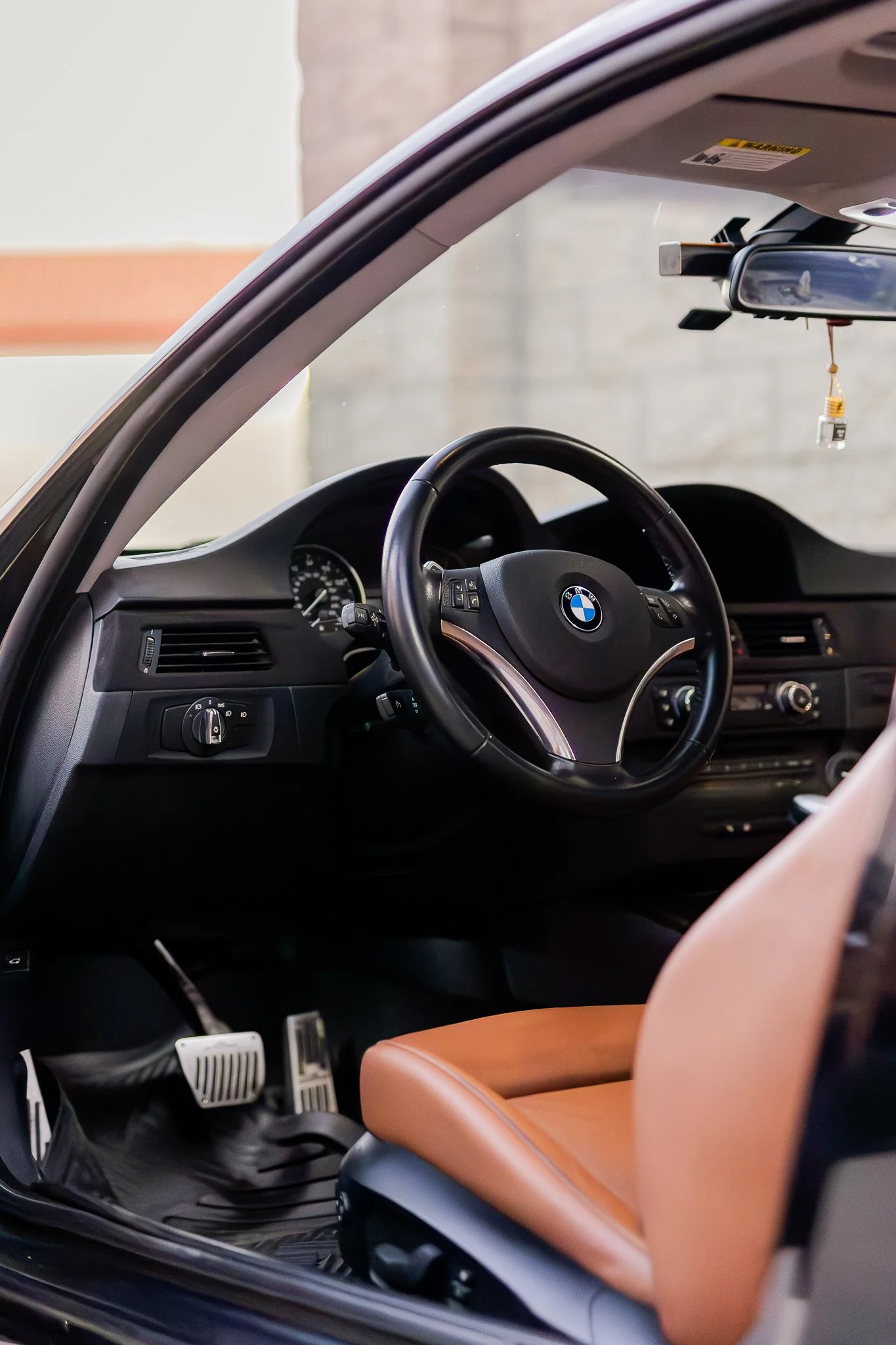 Interior of a BMW car showing the steering wheel, dashboard, brown leather seat, rearview mirror, and part of the front passenger seat.