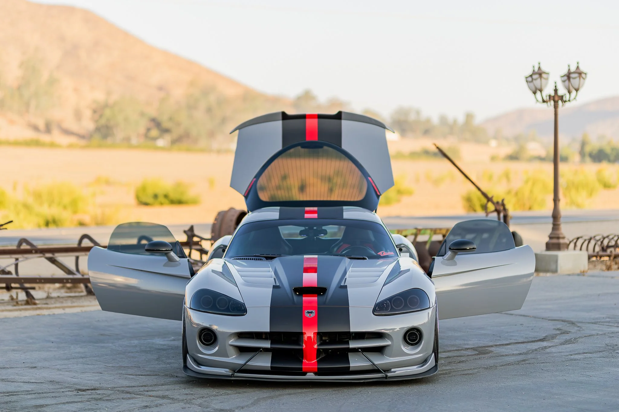 A silver sports car with red racing stripes and its gull-wing doors open, parked in an outdoor area with vintage lampposts and a scenic mountain background.