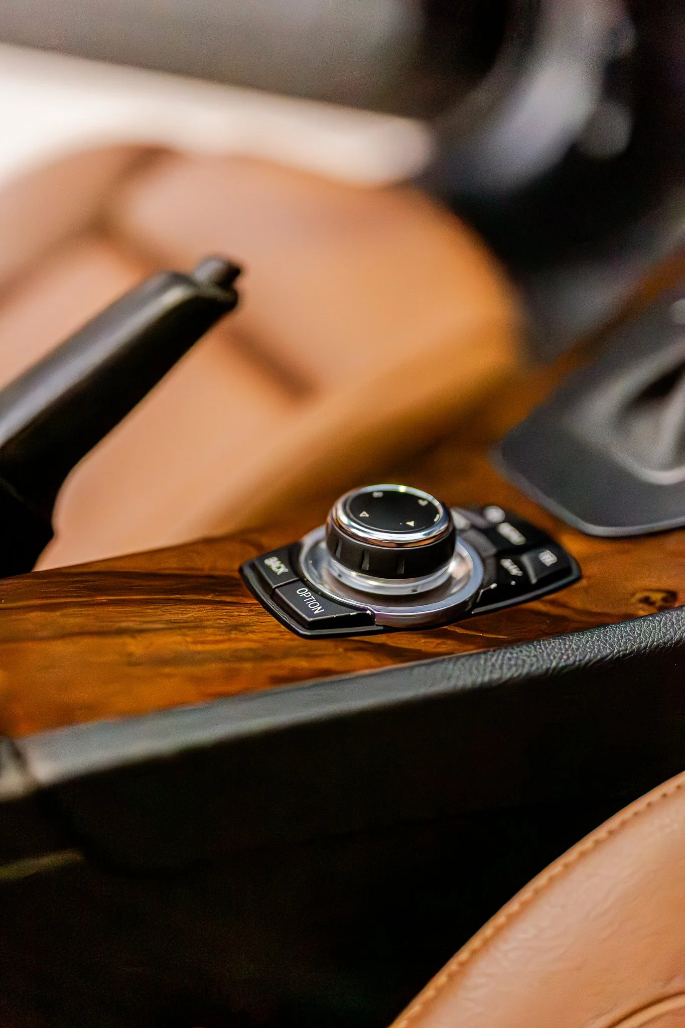 Close-up of a car's center console with a brown leather seat and control knob. Hand wearing a watch is seen resting nearby.