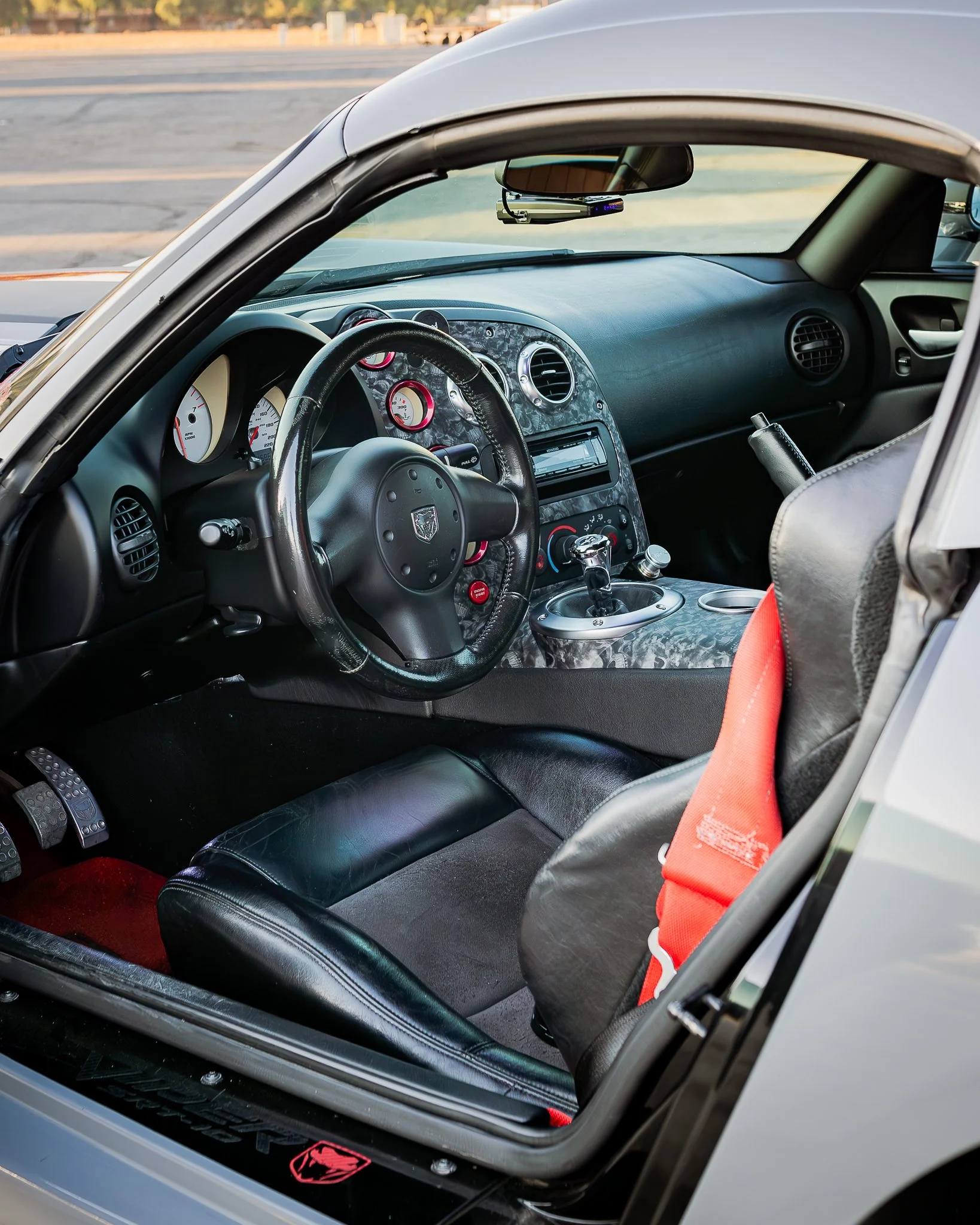 Interior of a car showing black leather seats with red accents, a dashboard with multiple gauges, and a racing style gear shifter. The steering wheel has a Dodge logo, and the rearview mirror is visible. The outside view shows a parking lot with some trees and a fence.