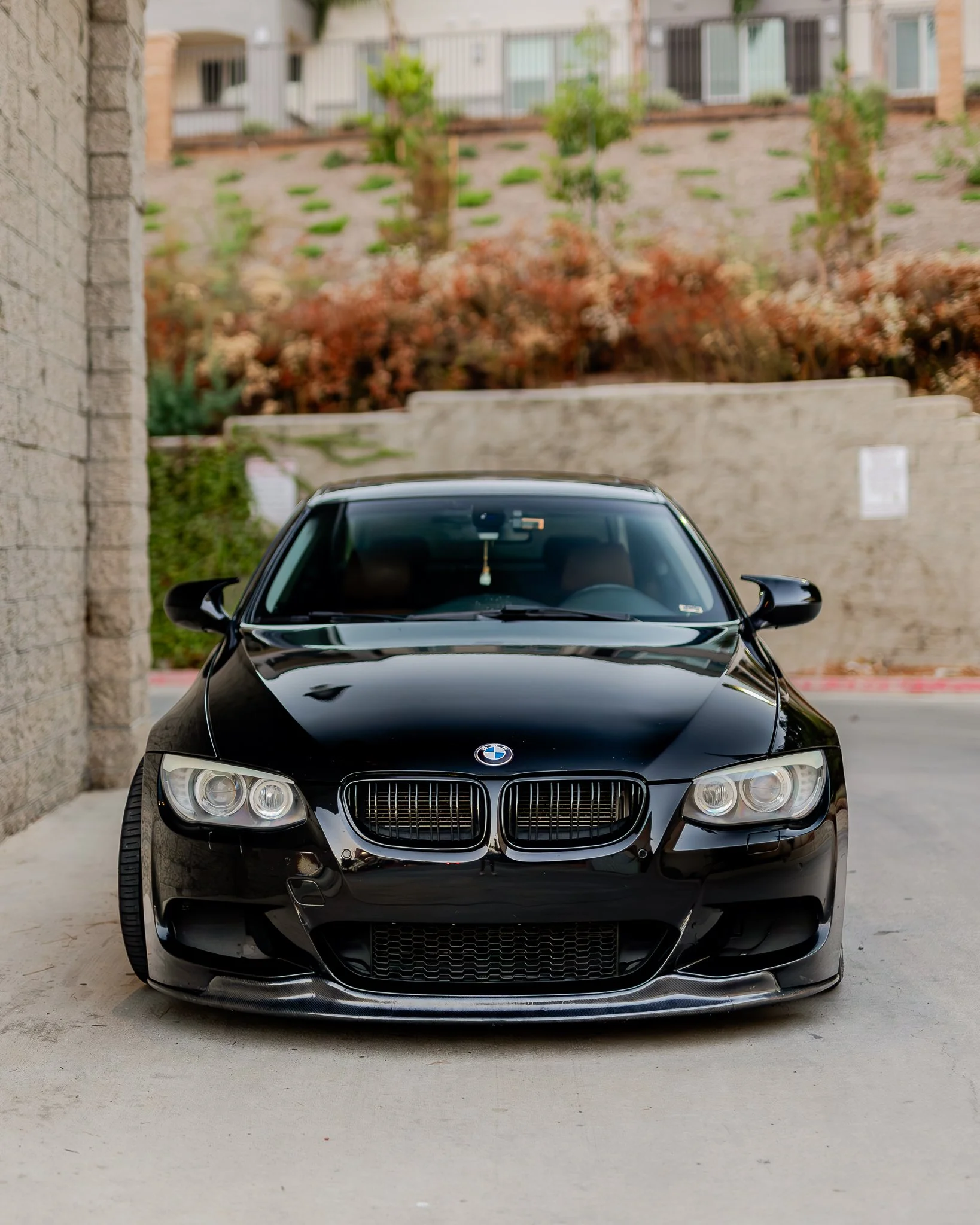Front view of a black BMW car parked outdoors on a concrete surface with a beige brick wall on the left and a stone wall with plants and murder in the background.