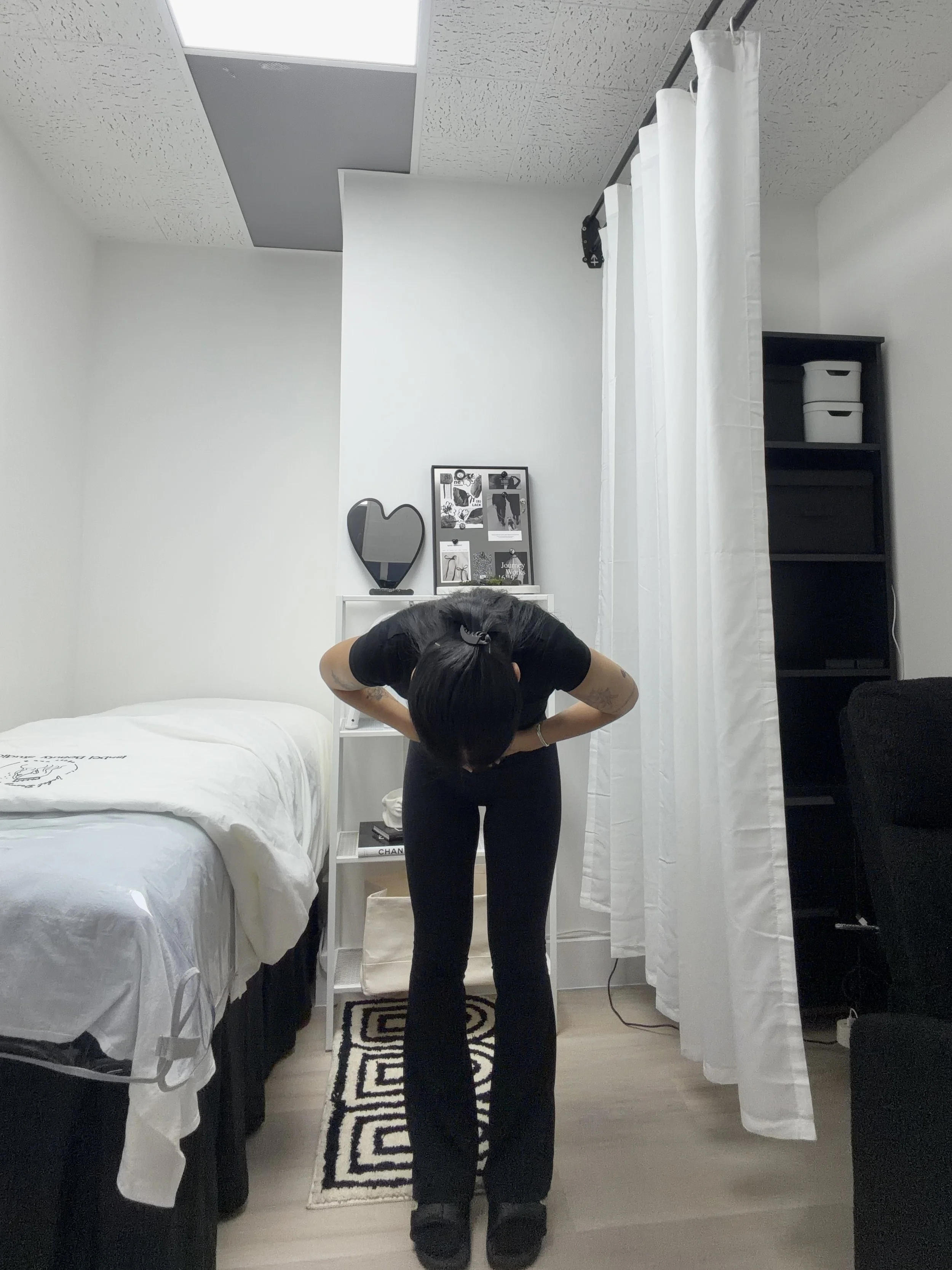 Person in black workout clothes bending forward in a bedroom beside a bed, white curtains, and shelving.