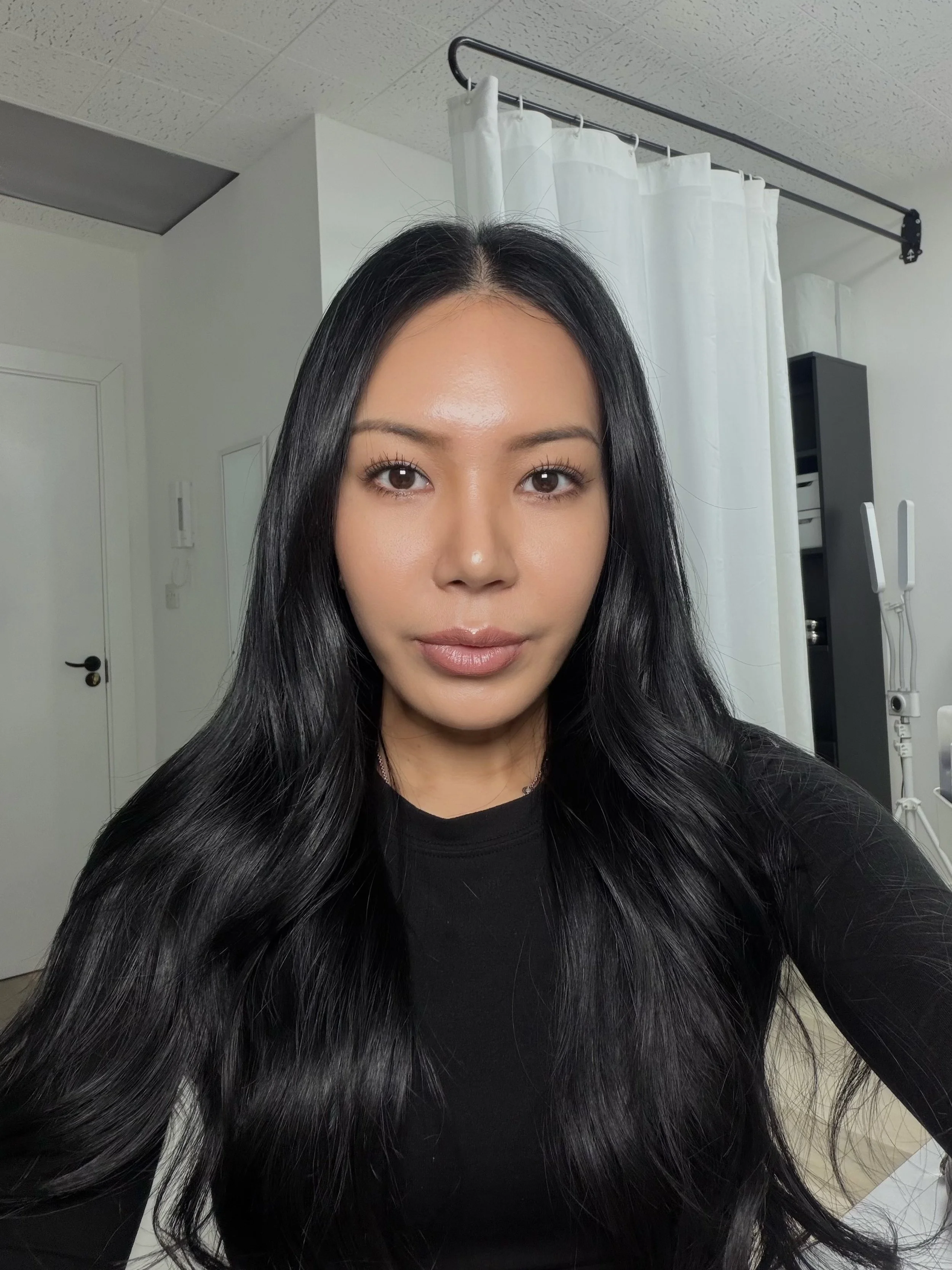 Close-up portrait of a woman with long black hair wearing a black shirt in a clinical room with privacy curtains.